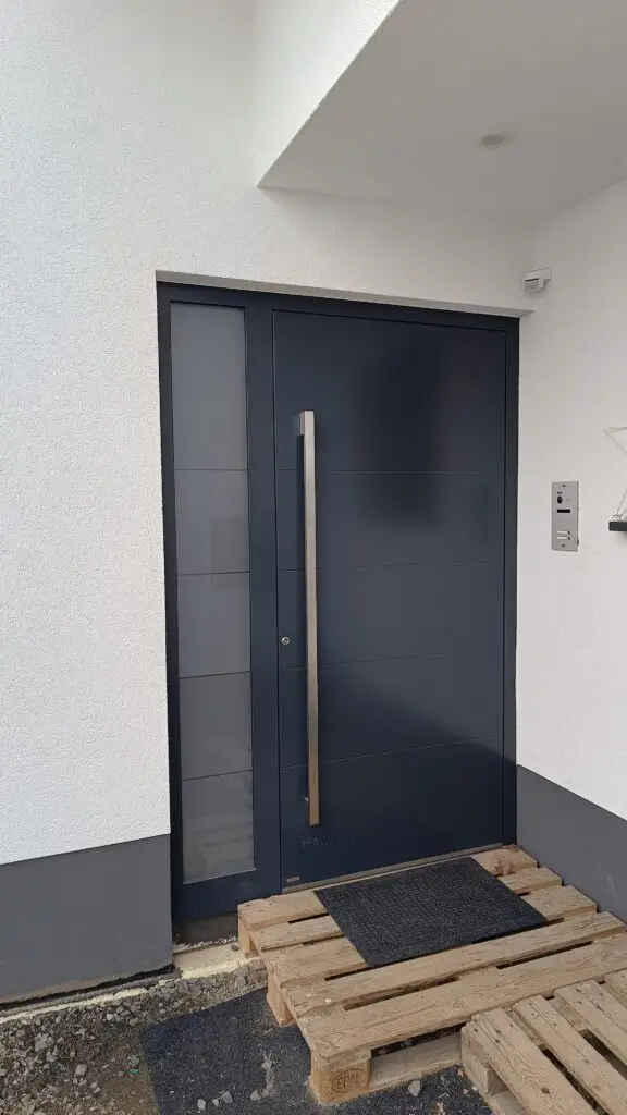 Anthrazitfarbene Haustür mit Edelstahl-Stangengriff, schmalem Glasseitenteil und Klingelanlage mit Kamera